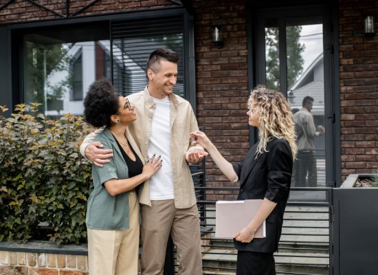 successful property agent giving key to happy multiethnic couple near house on city street