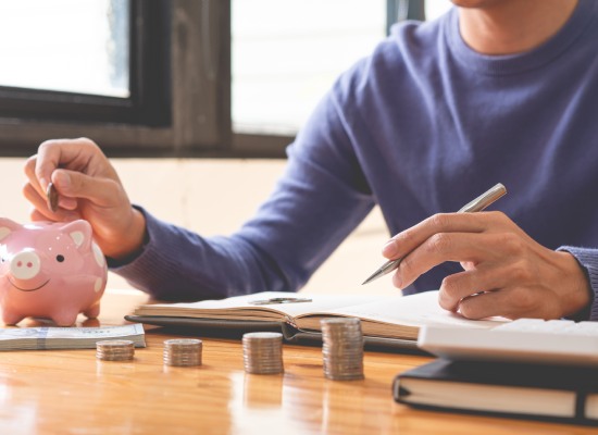 Man writing down his income and counting some money as a saving-up part on his notebook while dropping a coin into the piggy bank.