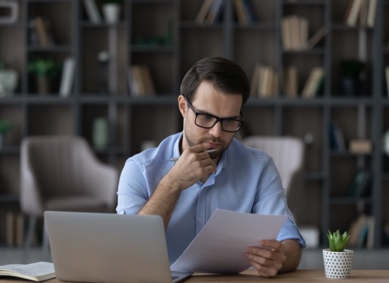 Working with papers. Serious millennial male entrepreneur work at home office alone hold read document hardcopy think on conditions terms. Focused young man engaged in paperwork edit text of agreement