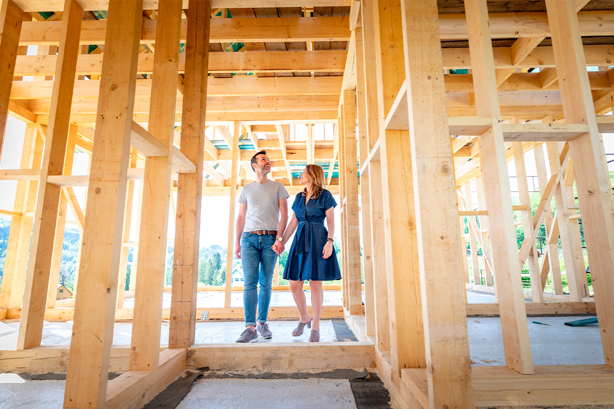 A couple poses inside a wooden frame under construction, representing their plans to buy land and build a house.